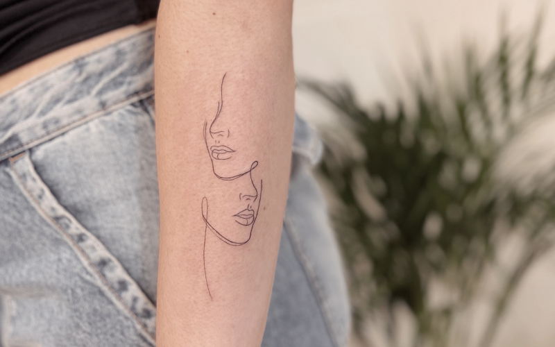 Fineline tattoo van twee gezichten in één lijn op de onderarm.