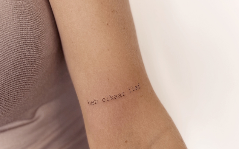 Minimalistische 'heb elkaar lief' tekst tattoo op de binnenarm.