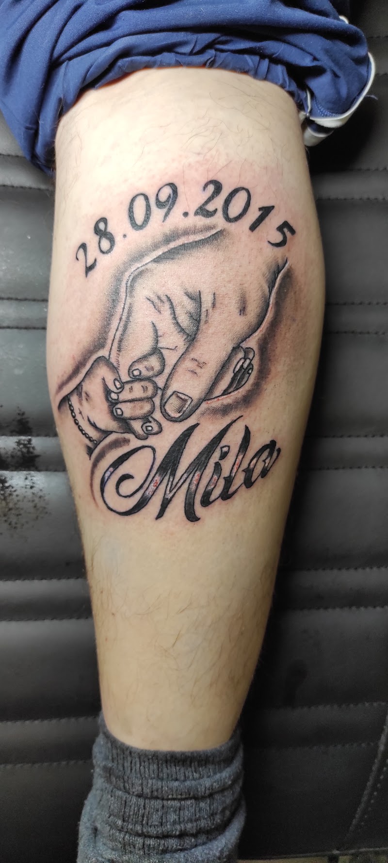 Blackwork tattoo van een volwassen hand die een babyhandje vasthoudt, met naam en datum.