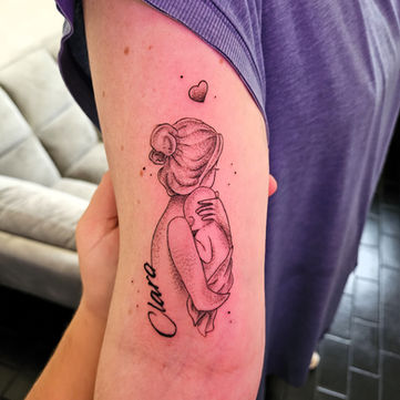 Fineline tattoo van moeder met baby en naam 'Clara' op bovenarm.