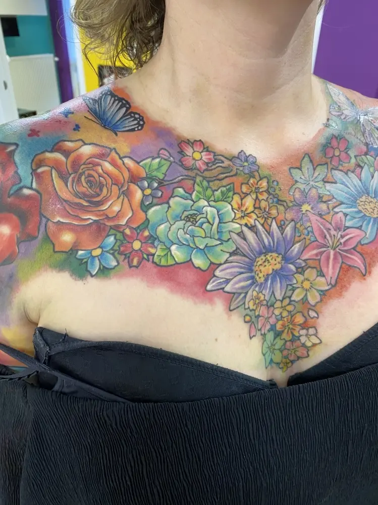 Kleurrijke bloemen en vlinder tattoo op borst en schouders.