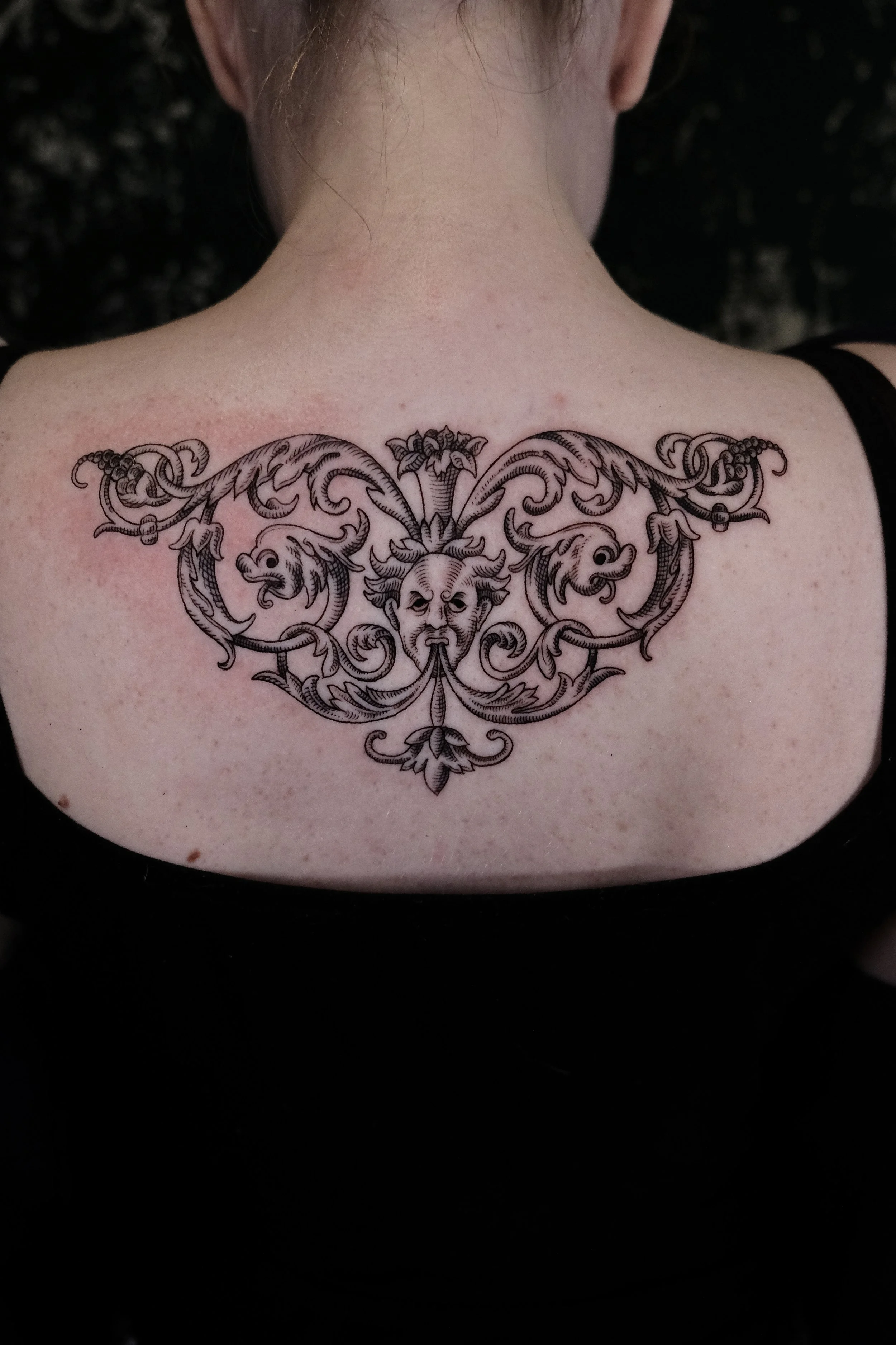Gedetailleerde blackwork tattoo van een grotesk gezicht met sierlijke krullen op de bovenrug.