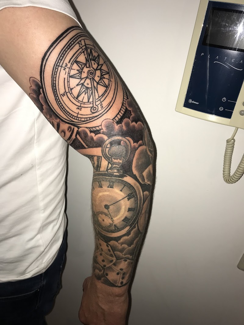 Zwart-wit tattoo van kompas, zakhorloge en dobbelstenen op arm.
