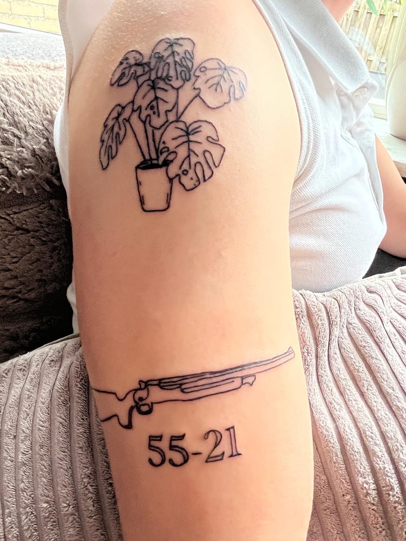 Fineline tattoos van een Monstera plant en een geweer met nummers op de bovenarm.