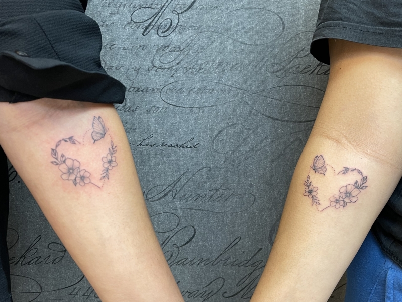 Twee identieke fineline tattoos van een hart gevormd door bloemen en een vlinder.